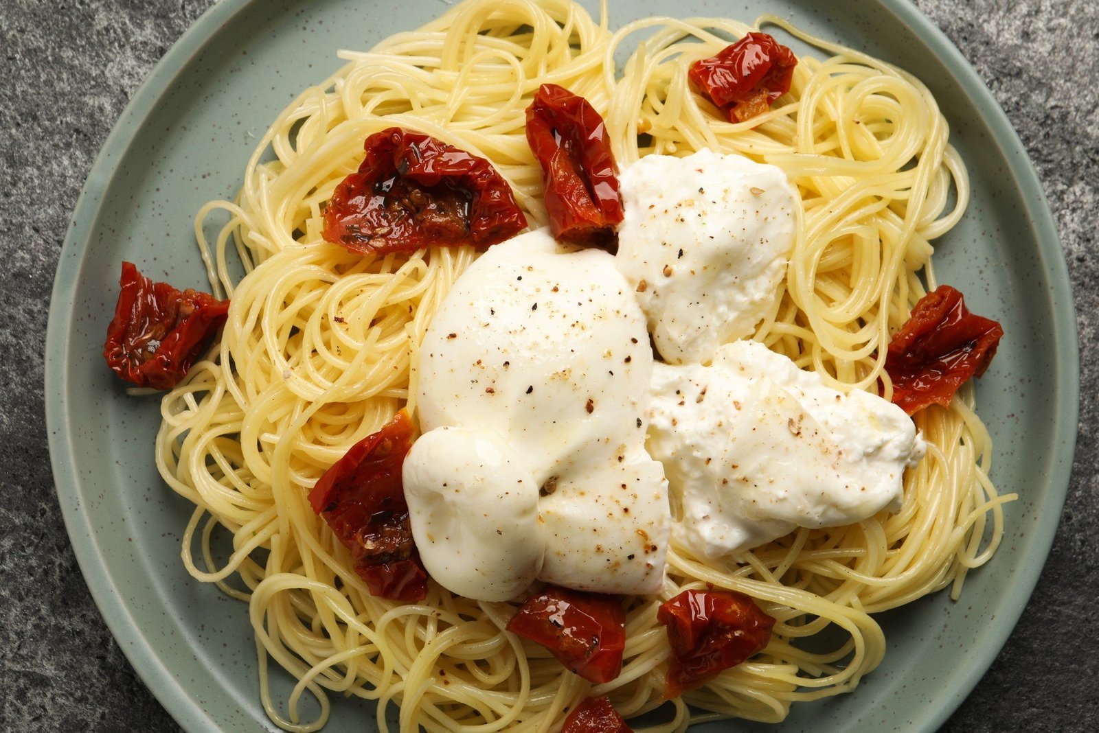 Spaghetti burrata pomodori grigliati serviti in un piatto bianco con foglie di basilico fresco e pepe nero macinato.